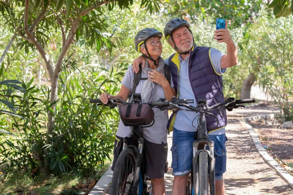 Active caucasian senior couple with electrobikes standing outdoors in the park video calling by cellphone. Two smiling elderly people wearing helmet running in nature enjoying healthy lifestyle. E-bike tours for seniors