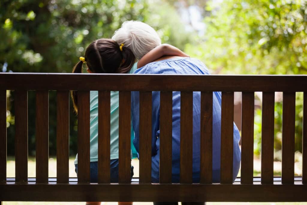 Rear view of granddaughter with arm around grandmother sitting on wooden bench at backyard. Lead photo for the blog post 11 Amazing Tips for Traveling with Grandchildren