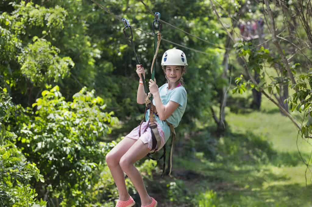 Happy smiling little girl riding a zip line in a lush tropical forest while on a vacation with grandparents