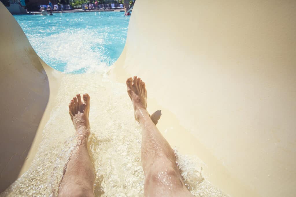 Point-of-view photo of a man going down a waterslide at an outdoor waterpark during a warm summer day. Focus on the feet and water slide. Cover photo of the post, "Kidult Vacations for Retirees"