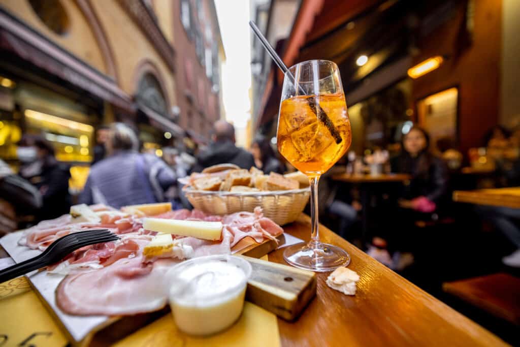 Meat plate with sliced italian sausages, proscutto, cheese, bread and Spritz Aperol drink on a table at outdoor bar on crowded street background. Part of every great foodie tour