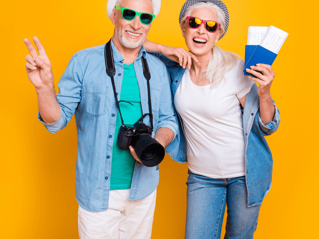 active older couple getting to enjoy a hobby that lets them travel the world. He's holding a camera, she's holding plane tickets.