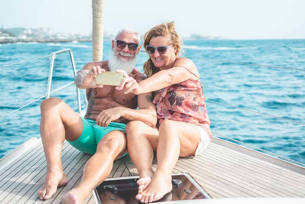 Senior couple doing selfie on sailboat during luxury ocean trip vacation - Old trendy people having fun on exclusive holiday - Travel and joyful elderly concept - Focus on faces