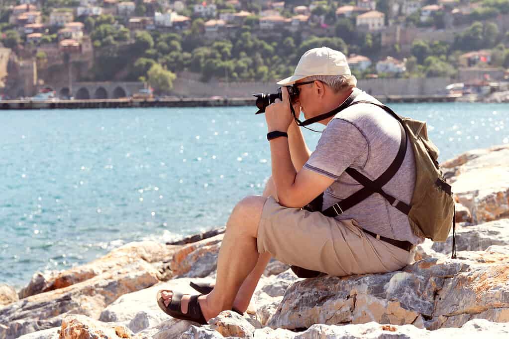 Older Man with camera sitting on the rocks on the shore. Tourist taking photo of sea against maritime city. One of many great hobbies for retired men