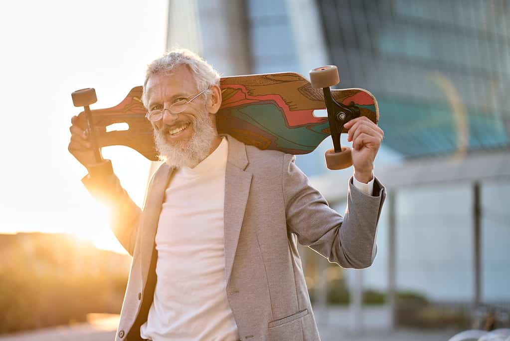 Smiling happy cool grey haired bearded older senior business man skater wearing suit holding skateboard standing in city on sunset outdoors after work. Old people freedom spirit concept. Portrait. healthy habits for reitrees