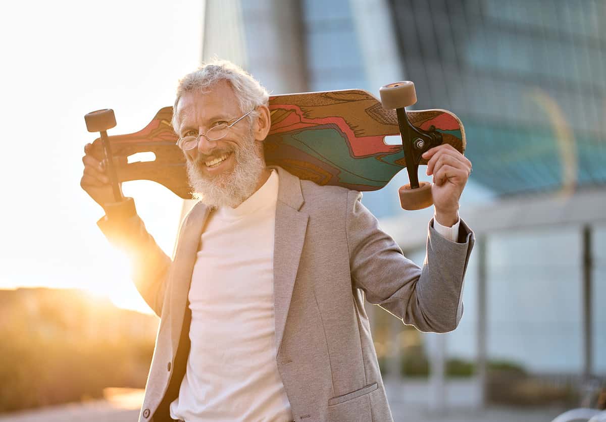 Smiling happy cool grey haired bearded older senior business man skater wearing suit holding skateboard standing in city on sunset outdoors after work. Old people freedom spirit concept. Portrait. healthy habits for reitrees