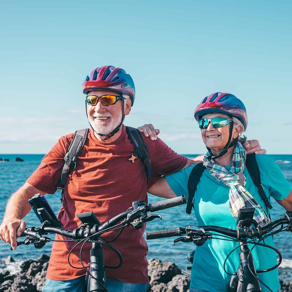 Two happy senior people enjoying riding bikes together at sea to be fit and healthy. Active senior couple wearing helmets having fun training in outdoors. Authentic elderly retired life concept