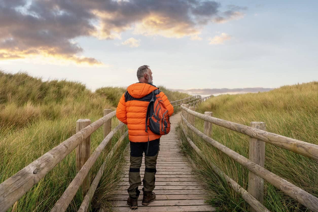 A man strolls on a wooden boardwalk surrounded by tall grass, heading toward a picturesque sunset backdrop and enjoying nature's beauty. Atelics for retirement, the new midlife crisis